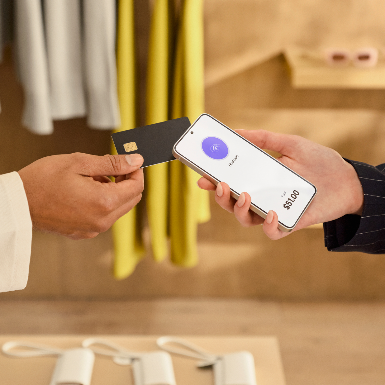 Two people are standing in a large, warm retail environment. The customer on the left holds a credit card to tap the sales associate’s Android, and the screen says “Hold card”. They background features wooden shelves with neatly stacked items, creating a warm, organized setting.